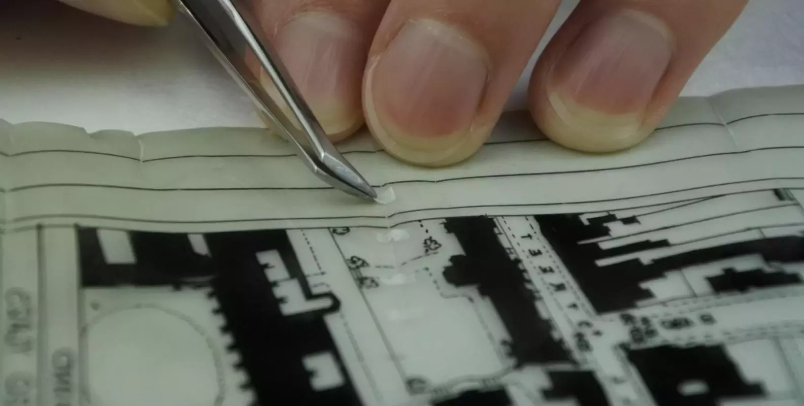 Close-up photograph of a small splint repair of Japanese kozo tissue being applied with tweezers to an architectural plan on plastic film.